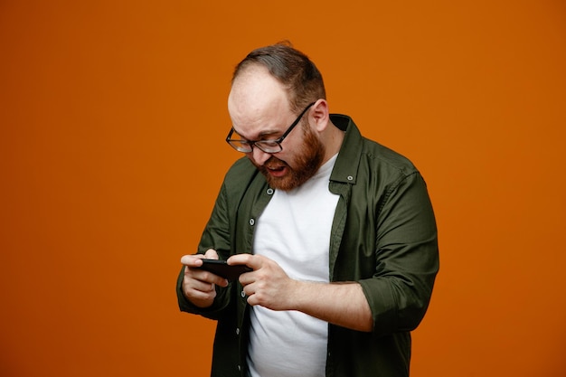 Uomo barbuto in abiti casual che indossano occhiali con in mano lo smartphone guardandolo confuso e deluso in piedi su sfondo arancione