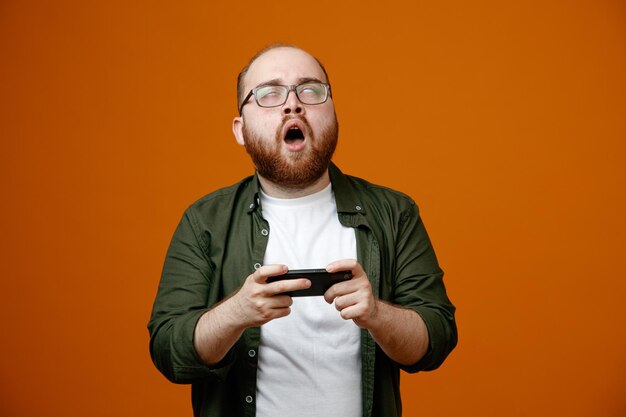 Uomo barbuto in abiti casual che indossano occhiali con smartphone che giocano a giochi che sembrano delusi in piedi su sfondo arancione