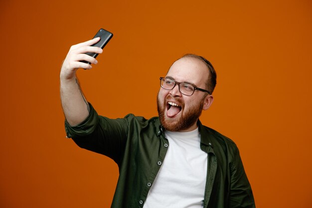 Uomo barbuto in abiti casual che indossano occhiali facendo selfie utilizzando smartphone felice e gioioso sporgendo lingua in piedi su sfondo arancione