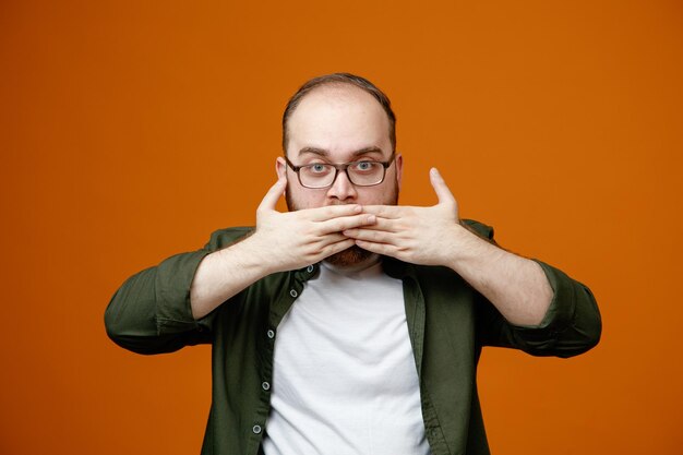 Uomo barbuto in abiti casual che indossano occhiali guardando la fotocamera con il viso serio che copre la bocca con le mani in piedi su sfondo arancione