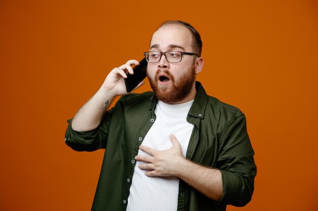 Uomo barbuto in abiti casual con gli occhiali che sembra confuso mentre parla al telefono cellulare in piedi su sfondo arancione