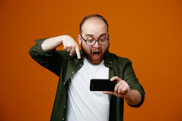Uomo barbuto in abiti casual con occhiali che tengono lo smartphone puntato con il dito indice guardandolo stupito e sorpreso in piedi su sfondo arancione