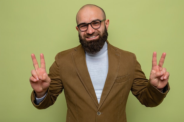 Uomo barbuto in abito marrone con gli occhiali che sembra sorridente allegramente mostrando il v-sign