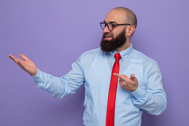 Uomo barbuto in cravatta rossa e camicia blu con gli occhiali che guarda da parte felice e contento che si presenta con il braccio della mano che punta con il dito indice a lato