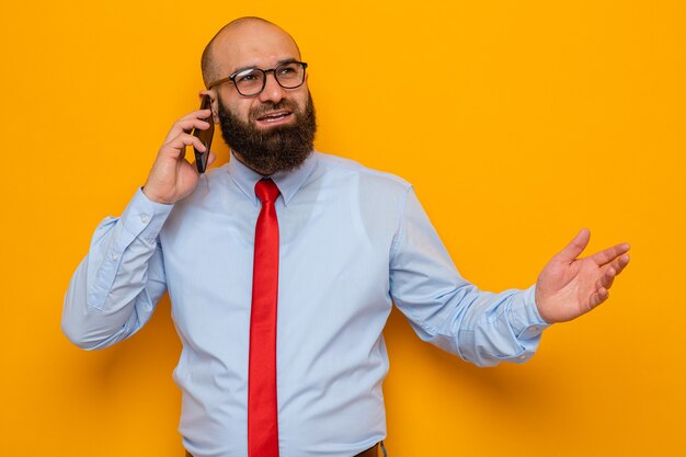 Uomo barbuto in cravatta rossa e camicia blu con gli occhiali che guarda da parte felice e positivo sorride allegramente mentre parla al telefono cellulare