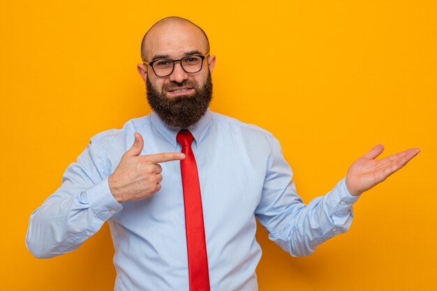 Uomo barbuto in cravatta rossa e camicia blu con gli occhiali che si presenta con il braccio della mano che punta con il dito indice di lato