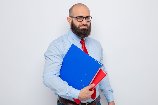 Uomo barbuto in cravatta rossa e camicia blu con gli occhiali che tengono le cartelle dell'ufficio guardando la telecamera con un'espressione seria sicura in piedi su sfondo bianco