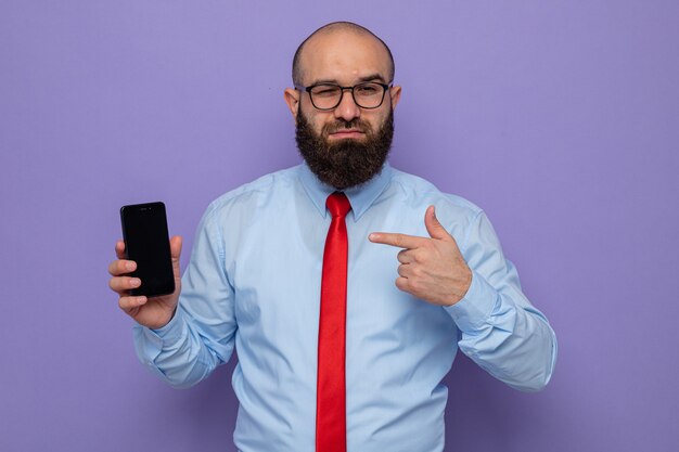 Uomo barbuto in cravatta rossa e camicia blu con gli occhiali che tengono lo smartphone puntato con il dito indice e sembra sorridente sicuro confident