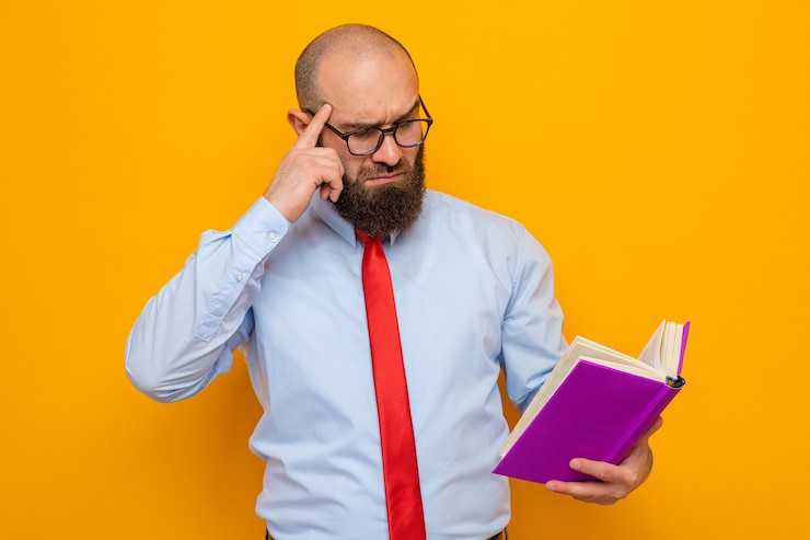 Foto gratuito uomo barbuto in cravatta rossa e camicia blu con gli occhiali che tiene un libro guardandolo pensando perplesso