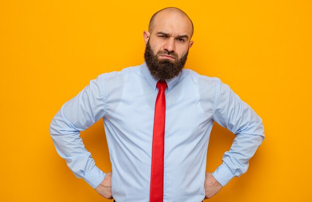 Uomo barbuto in cravatta rossa e camicia che guarda l'obbiettivo con espressione scettica sul viso in piedi su sfondo arancione