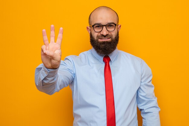 Uomo barbuto in cravatta rossa e camicia con gli occhiali guardando la telecamera sorridente fiducioso che mostra il numero tre con le dita in piedi su sfondo arancione