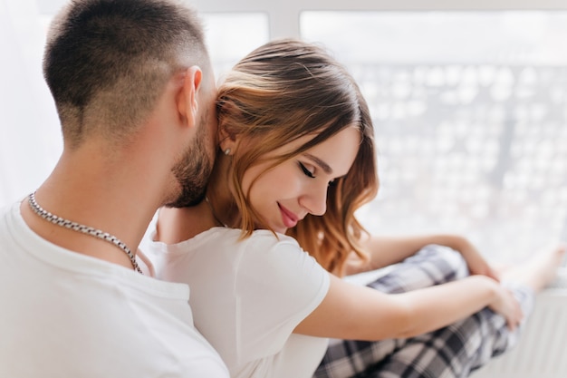 Uomo barbuto in maglietta bianca che bacia i capelli della ragazza durante il servizio fotografico in interni. Ritratto di donna bionda riccia accattivante che abbraccia con il marito.