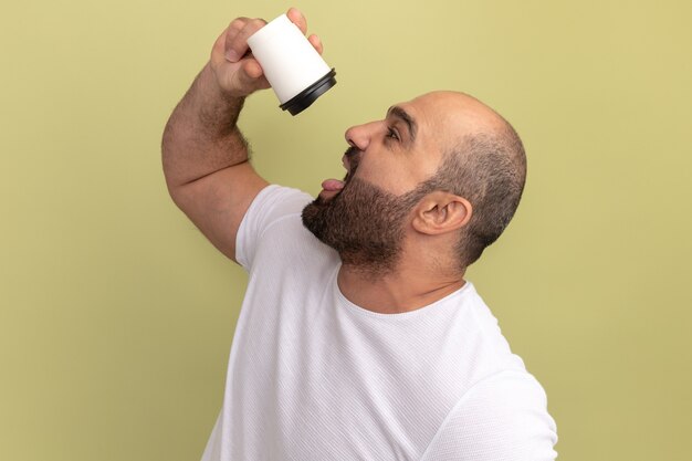 Uomo barbuto in maglietta bianca che tiene tazza di caffè andando a bere in piedi sopra la parete verde