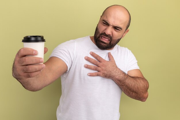 Uomo barbuto in maglietta bianca che tiene tazza di caffè guardandolo con espressione disgustata in piedi sopra la parete verde