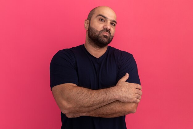 Uomo barbuto in maglietta blu scuro che osserva da parte dispiaciuto con le braccia incrociate in piedi sopra il muro rosa