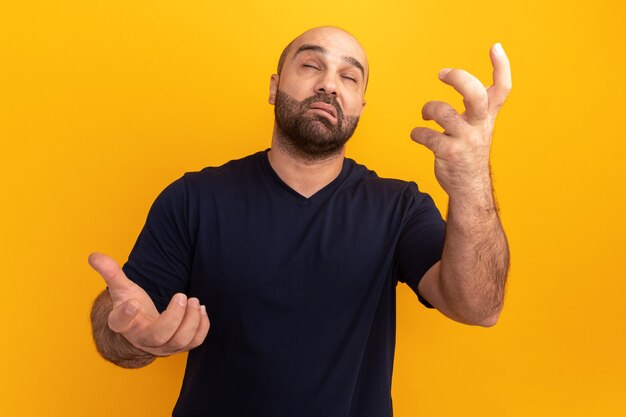 Uomo barbuto in maglietta blu scuro che sembra confuso gesti con le mani in piedi sopra la parete arancione