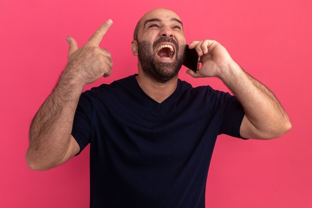 Uomo barbuto in maglietta blu scuro che sembra gridare infastidito impazzire mentre parla al telefono cellulare in piedi sopra il muro rosa