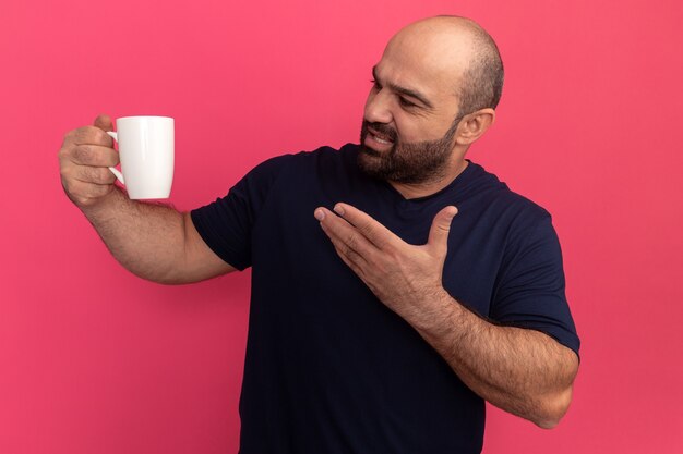 Uomo barbuto in maglietta blu scuro che tiene una tazza che punta con il braccio alla tazza con espressione infastidita in piedi sopra il muro rosa