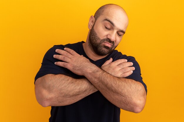 Uomo barbuto in maglietta blu scuro con gli occhi chiusi con le braccia incrociate sul petto in piedi sopra il muro arancione