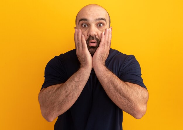 Uomo barbuto in maglietta blu scuro stupito e sorpreso in piedi sopra la parete arancione