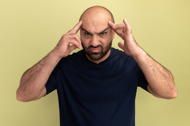 Uomo barbuto in maglietta nera che sembra malato e infastidito toccando la sua testa che soffre di forte mal di testa in piedi sopra il muro verde