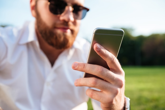 uomo barbuto in occhiali da sole e camicia mentre si utilizza lo smartphone. Focus sul telefono