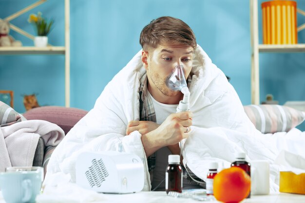 Uomo barbuto malato con canna fumaria seduto sul divano a casa coperto con una coperta calda e utilizzando un inalatore quando si tossisce.