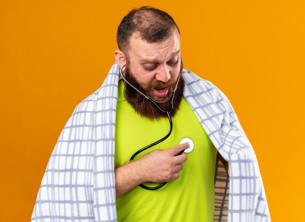 Uomo barbuto malsano avvolto in una coperta che si sente male soffre di freddo ascoltando il suo battito cardiaco usando lo stetoscopio essendo preoccupato in piedi sul muro arancione