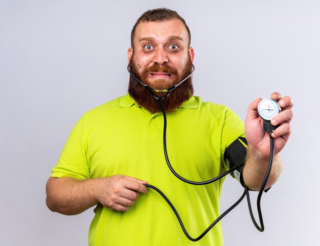 Uomo barbuto malsano in polo gialla che si sente male misurando la pressione sanguigna usando il tonometro che sembra preoccupato in piedi sul muro bianco