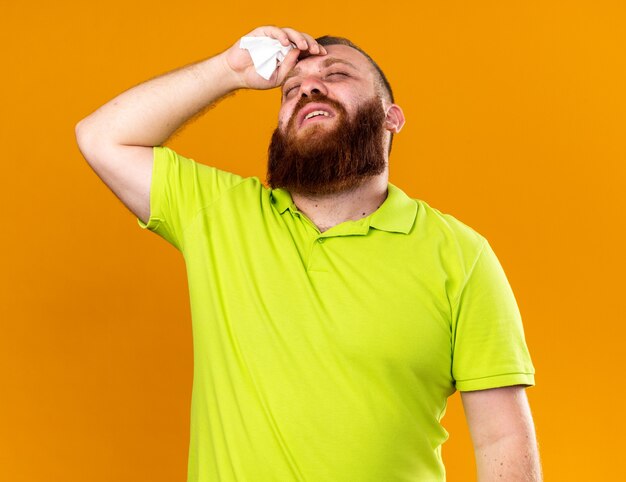 Uomo barbuto malsano in polo gialla che si sente terribilmente soffre di freddo con febbre con la mano sulla fronte in piedi sul muro arancione