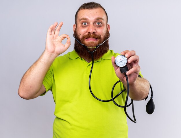 Uomo barbuto malsano in polo gialla sentirsi male misurando la pressione sanguigna usando il tonometro confuso mostrando segno ok in piedi sul muro bianco