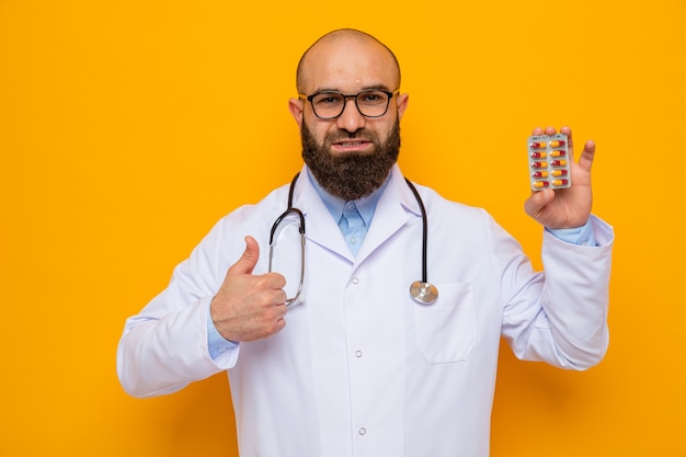 Uomo barbuto medico in camice bianco con stetoscopio intorno al collo con gli occhiali che tengono blister con pillole guardando la telecamera sorridendo allegramente mostrando i pollici in piedi su sfondo arancione