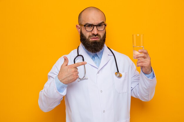 Uomo barbuto medico in camice bianco con uno stetoscopio intorno al collo con gli occhiali tenendo un bicchiere di acqua puntando con il dito indice contro di esso con faccia grave