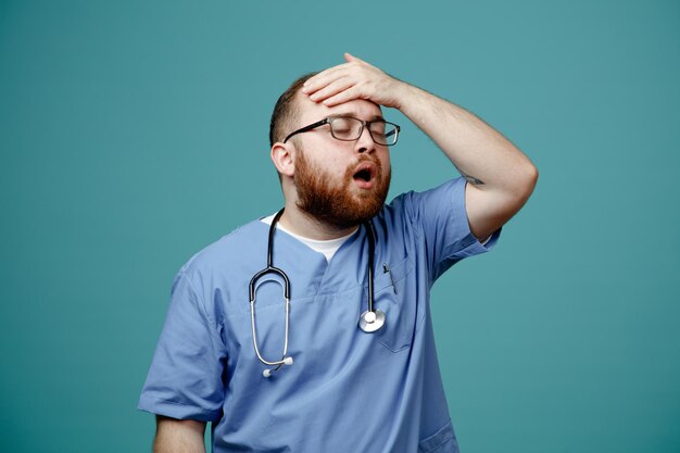 Uomo barbuto medico in uniforme con stetoscopio intorno al collo con gli occhiali che sembra confuso e deluso tenendo la mano sulla testa per errore in piedi su sfondo blu