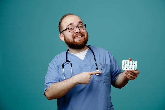 Uomo barbuto medico in uniforme con uno stetoscopio intorno al collo che indossa occhiali con in mano le pillole che puntano con il dito indice verso le pillole sorridenti con la faccia felice in piedi su sfondo blu