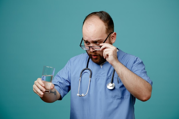 Uomo barbuto medico in uniforme con uno stetoscopio intorno al collo che indossa occhiali con in mano un bicchiere d'acqua guardandolo da vicino con la faccia seria in piedi su sfondo blu