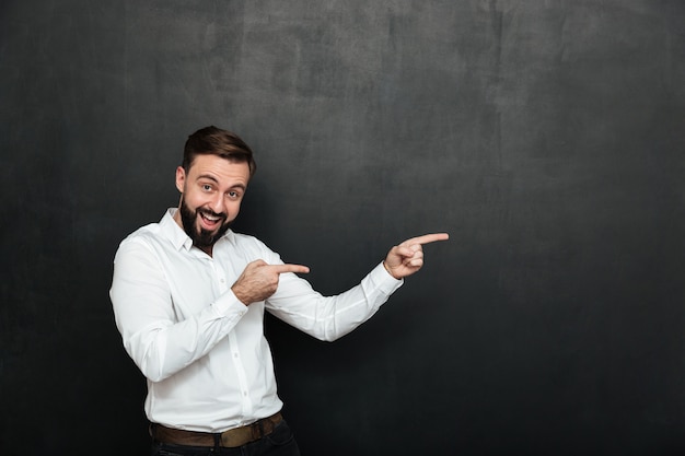 Uomo barbuto ottimista in camicia bianca che indica i dito indice da parte, dimostrando o facendo pubblicità sopra lo spazio grigio scuro della copia