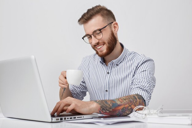 Uomo barbuto positivo con pettinatura alla moda, indossa una camicia formale, beve cappuccino o caffè espresso,