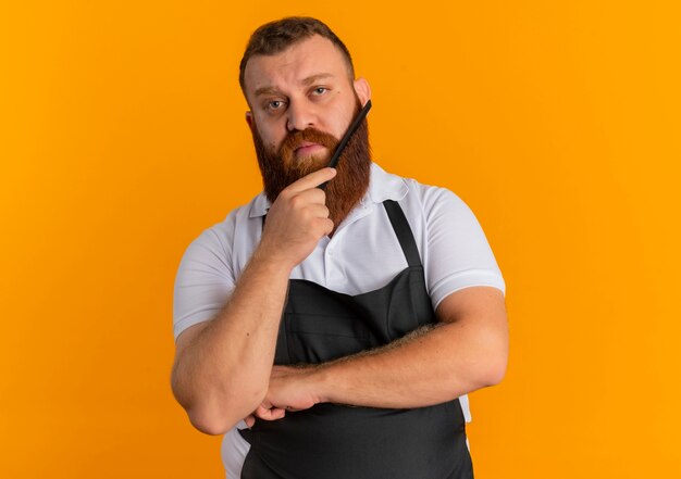 Uomo barbuto professionale del barbiere in grembiule che pettina la sua barba con espressione fiduciosa in piedi sopra la parete arancione