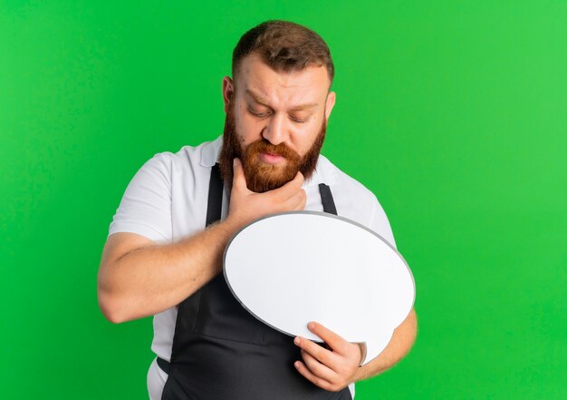 Uomo barbuto professionale del barbiere in grembiule che tiene il segno in bianco del fumetto perplesso con espressione pensierosa che sta sopra la parete verde