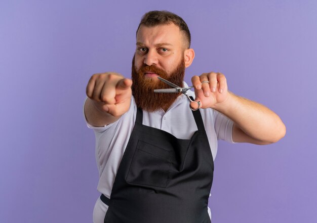 Uomo barbuto professionale del barbiere in grembiule che tiene le forbici che indicano con il dito indice dispiaciuto in piedi sopra la parete viola