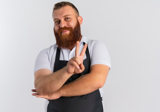 Uomo barbuto professionista del barbiere in grembiule che sorride allegramente mostrando vittoria cantare in piedi sopra il muro bianco