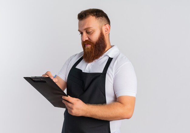 Uomo barbuto professionista del barbiere in grembiule che tiene appunti guardandolo con faccia seria in piedi sopra il muro bianco