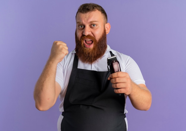 Uomo barbuto professionista del barbiere in grembiule che tiene macchina per il taglio dei capelli stringendo il pugno con espressione aggressiva in piedi sopra la parete viola