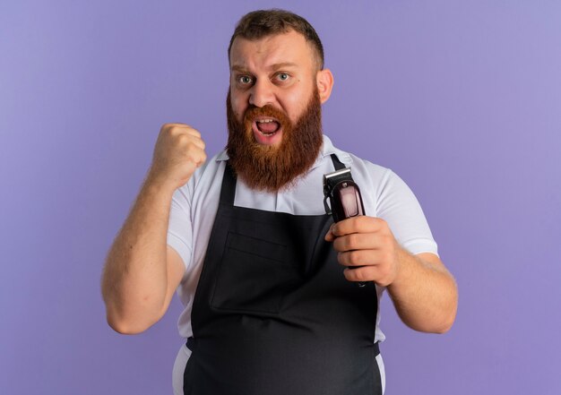 Uomo barbuto professionista del barbiere in grembiule che tiene macchina per il taglio dei capelli stringendo il pugno con espressione aggressiva in piedi sopra la parete viola