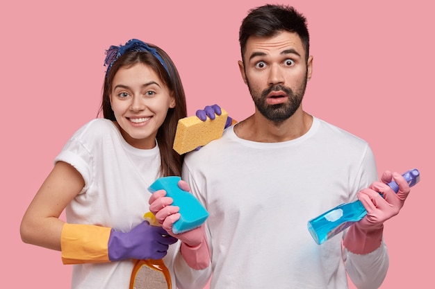 Uomo barbuto scioccato, positivo giovane donna graziosa usa prodotti per la pulizia per riordinare la stanza, fa i lavori domestici durante il giorno libero