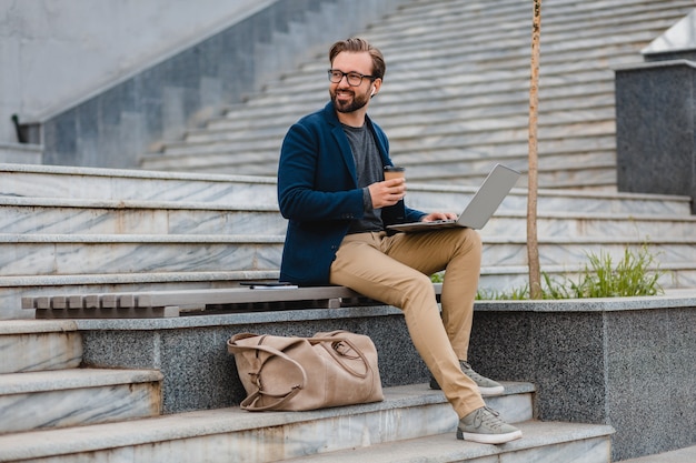 Uomo barbuto sorridente bello in vetri che lavora al computer portatile