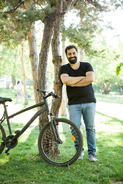 Uomo barbuto sorridente che si appoggia su un albero nel parco accanto alla sua bicicletta