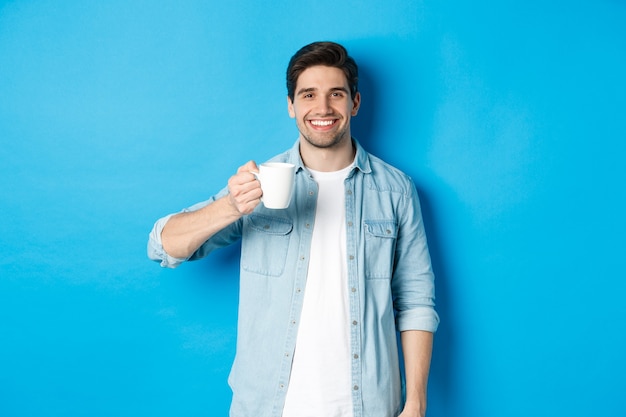 Uomo barbuto sorridente che tiene tazza e beve caffè, in piedi su sfondo blu.