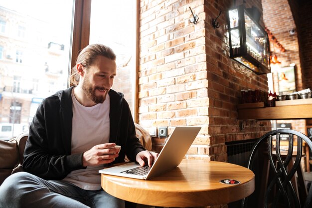 Uomo barbuto sorridente con il computer portatile in caffè